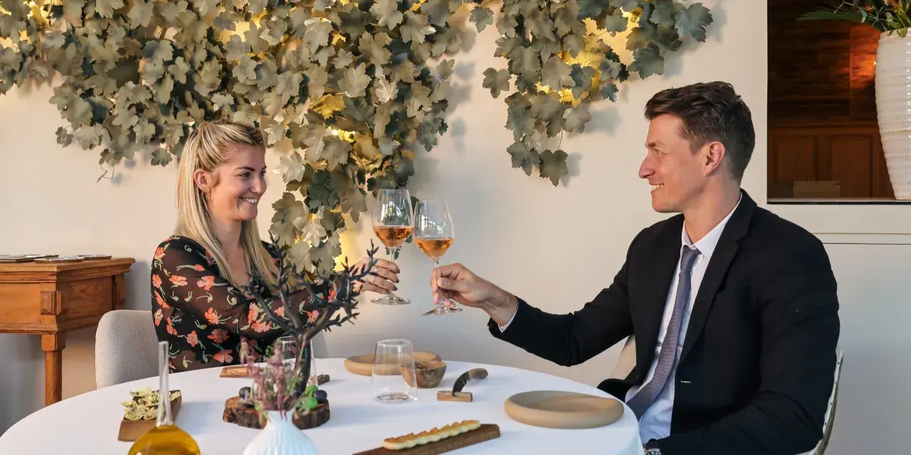 Un couple trinque avec du vin rosé dans un restaurant gastronomique, décoré de feuilles stylisées et d'une lampe design.