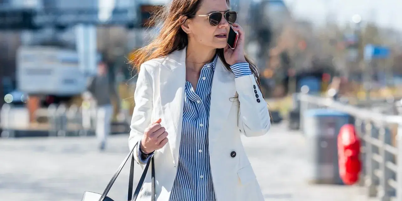 Une femme élégante porte une tenue avec blazer blanc, chemise à rayures bleues et un sac noir, tout en téléphonant.