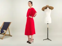 Une femme pose dans une robe rouge éclatante. À ses côtés, une chaise longue, un mannequin de couture et des plantes. Une robe casual, c'est quoi ?