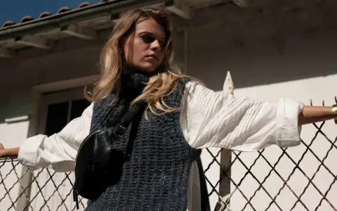 Une jeune femme porte un pull sans manche en maille bleue sur une chemise blanche, un sac banane noir et un bracelet doré. Elle s'appuie sur une clôture rouillée.