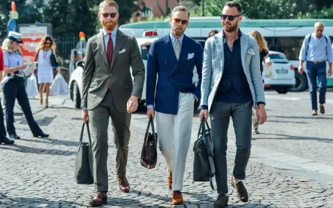 Trois hommes élégants déambulent. L'un porte une tenue avec blazer bleu ciel, un autre un costume vert olive, et le troisième un blazer gris clair.