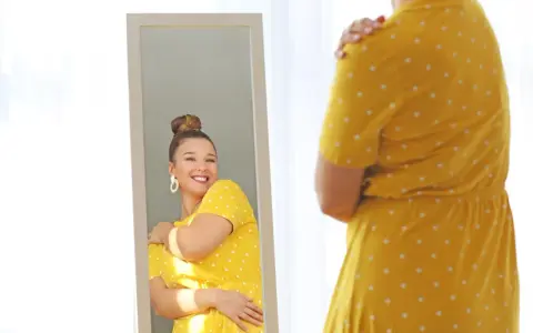 Une femme se regarde dans un miroir, souriante, portant une robe jaune à pois. Elle semble heureuse, malgré la robe trop courte.