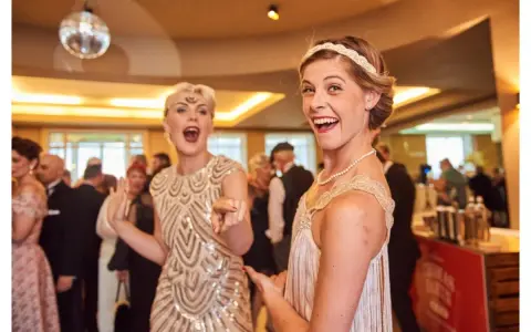 Deux femmes dansent, l'une porte une robe à paillettes dorées et un bandeau, l'autre une robe à franges, parfaites pour une tenue guinguette femme.