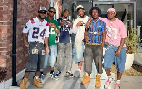 Six hommes posent ensemble, portant des tenues inspirées des années 2000.