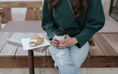 Une femme en pull vert, jean déchiré, savoure un café et un croissant. Parfait pour un look décontracté.