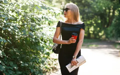 Une femme élégante, parfaite pour savoir comment porter robe noire à 50 ans, tient un café et un magazine, profitant d'une promenade ensoleillée.