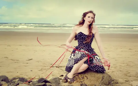 Jeune femme au style pin-up année 50, assise sur un rocher sur la plage, robe à pois, ruban rouge flottant au vent.