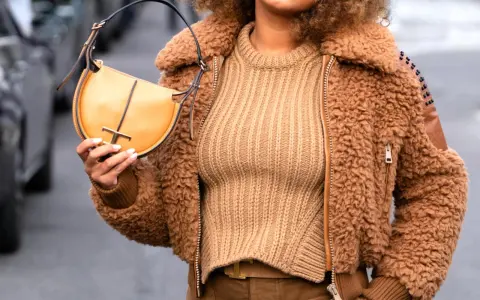 Une femme porte un pull beige côtelé, une veste en fausse fourrure marron et un sac à main jaune moutarde. Le jaune moutarde est une couleur qui va bien avec le beige.