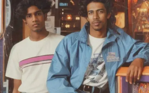 Deux jeunes hommes posent, arborant un style vestimentaire année 80 : t-shirt à rayures roses et veste bleue.