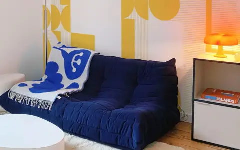 Canapé bleu profond avec plaid blanc et bleu, sur fond de papier peint jaune géométrique en **color blocking**. Lampe orange sur une étagère.