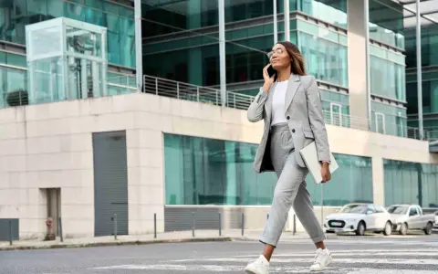 Femme en tenue d'entretien d'embauche d'été, tailleur gris, traversant la rue avec un ordinateur portable, parlant au téléphone.