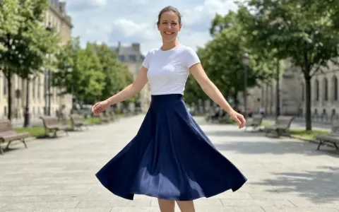 Une femme en talon et jupe évasée bleue danse joyeusement dans une rue parisienne bordée d'arbres.