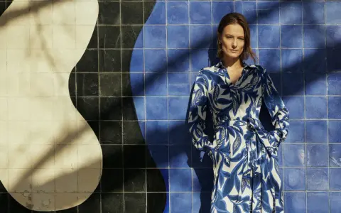 Une femme pose devant un mur de carreaux bleus et blancs. Sa robe à motifs bleus et blancs s'harmonise parfaitement avec le décor. Quelle couleur va bien avec le bleu vêtement ? Le blanc, évidemment !