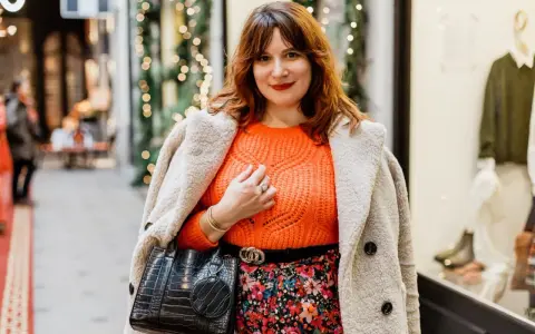 Une femme arbore une tenue avec pull orange vif, jupe fleurie et manteau douillet, prête pour une sortie festive.