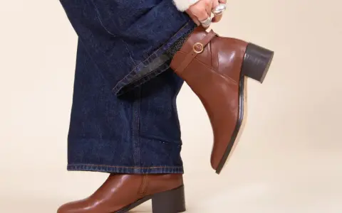 Une femme ajuste sa tenue avec bottines marron. Un jean évasé et un pull blanc complètent le look.