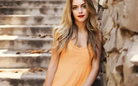 Jeune femme blonde, robe orange, appuyée contre un mur de pierres, escalier en arrière-plan.