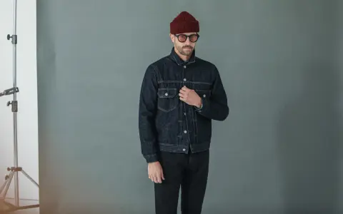 Homme portant une veste en jean sombre et un pantalon noir, bonnet bordeaux et lunettes rondes.
