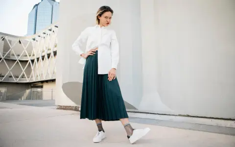 Une femme pose avec confiance, portant une chemise blanche et une élégante jupe plissée vert foncé.