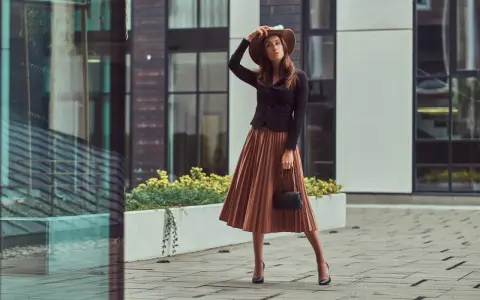 Une femme élégante porte une jupe longue plissée et des bottines à talons aiguilles, un chapeau à la main.