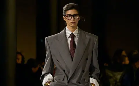 Mannequin au style collegienne, portant un costume gris oversize, chemise à rayures et cravate bordeaux. Lunettes imposantes.