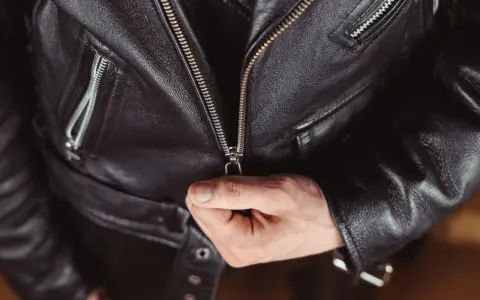 Main d'homme fermant la fermeture éclair d'une veste en cuir noire, partie d'un outfit classique et intemporel.