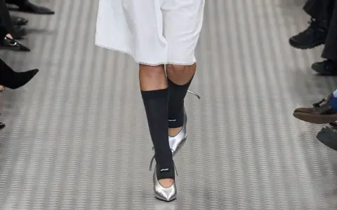 Une mannequin défile avec une tenue blanche et des chaussettes hautes grises, rehaussée par des chaussures argentées pointues.