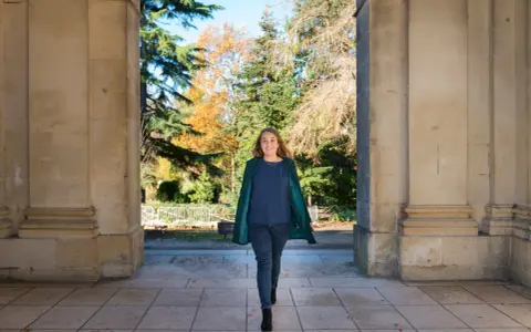 Une femme souriante marche sous une arche, portant un manteau vert et un haut bleu marine. L'automne colore les arbres en arrière-plan.