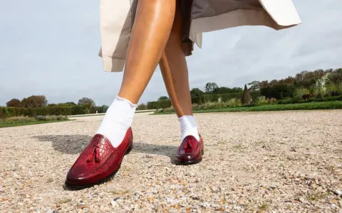 Pieds chaussés de mocassins rouges vernis, ornés de pompons, marchant sur un chemin de gravier. Le rouge vif de ces chaussures suggère audace et passion.