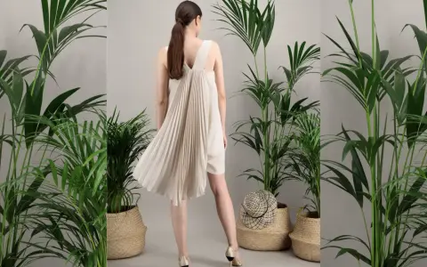 Une femme porte une robe plissée beige, parfaite pour une tenue mariage septembre. Elle est entourée de plantes vertes luxuriantes dans des paniers en osier.