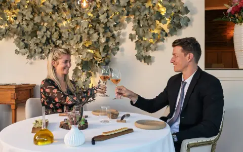 Un couple trinque avec du vin rosé dans un restaurant gastronomique, décoré de feuilles stylisées et d'une lampe design.