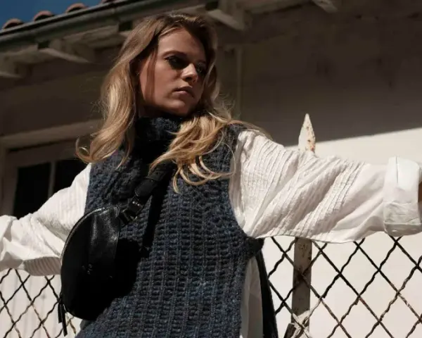 Une jeune femme porte un pull sans manche en maille bleue sur une chemise blanche, un sac banane noir et un bracelet doré. Elle s'appuie sur une clôture rouillée.