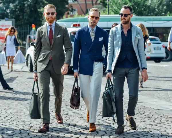 Trois hommes élégants déambulent. L'un porte une tenue avec blazer bleu ciel, un autre un costume vert olive, et le troisième un blazer gris clair.