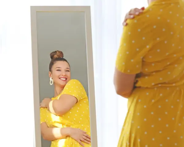 Une femme se regarde dans un miroir, souriante, portant une robe jaune à pois. Elle semble heureuse, malgré la robe trop courte.