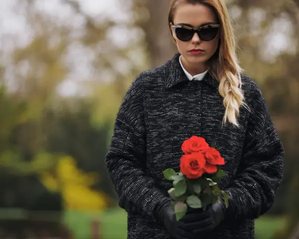 Une jeune femme, portant une tenue d'enterrement hiver, tient un bouquet de roses rouges. Ses lunettes de soleil cachent ses yeux, mais son expression est empreinte de tristesse.