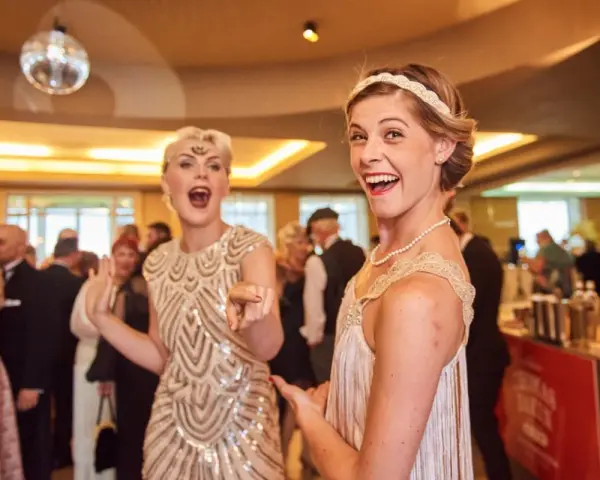 Deux femmes dansent, l'une porte une robe à paillettes dorées et un bandeau, l'autre une robe à franges, parfaites pour une tenue guinguette femme.