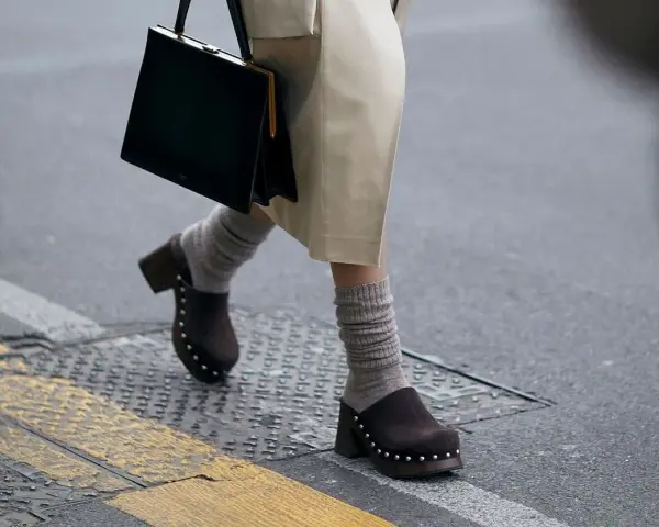 Une femme marche dans la rue, portant un sac noir et un élégant sabot femme marron clouté avec des chaussettes grises.