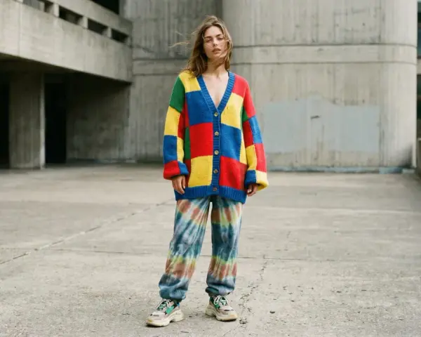 Jeune femme portant un gilet coloré style oversize et un pantalon tie-dye, devant un bâtiment en béton.