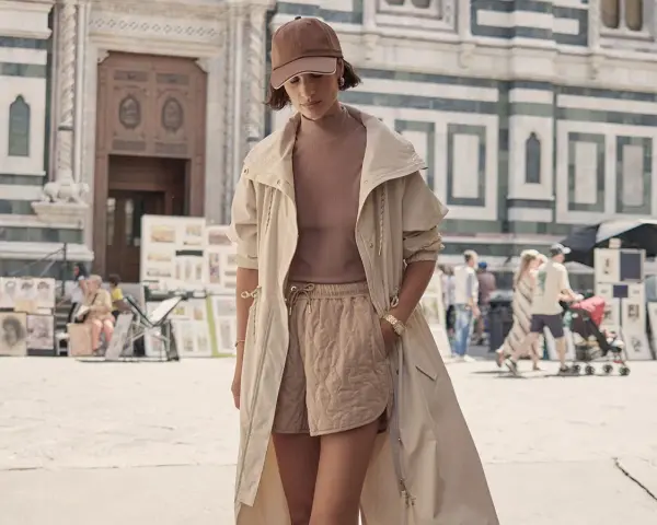 Une femme porte un ensemble athleisure chic : short matelassé, haut col roulé, veste longue et casquette. Elle se promène devant une cathédrale.