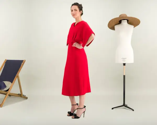 Une femme pose dans une robe rouge éclatante. À ses côtés, une chaise longue, un mannequin de couture et des plantes. Une robe casual, c'est quoi ?
