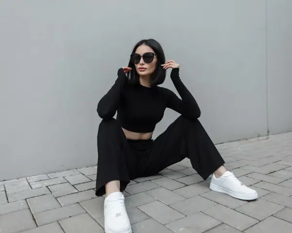 Une femme en tenue noire, crop top et pantalon large, assise sur des pavés. Idée de comment porter un pantalon large avec des baskets blanches.
