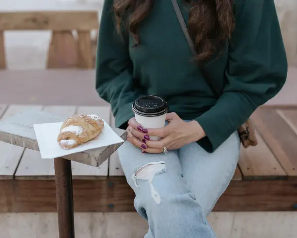 Une femme en pull vert, jean déchiré, savoure un café et un croissant. Parfait pour un look décontracté.