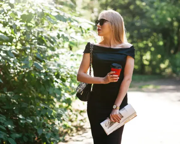 Une femme élégante, parfaite pour savoir comment porter robe noire à 50 ans, tient un café et un magazine, profitant d'une promenade ensoleillée.
