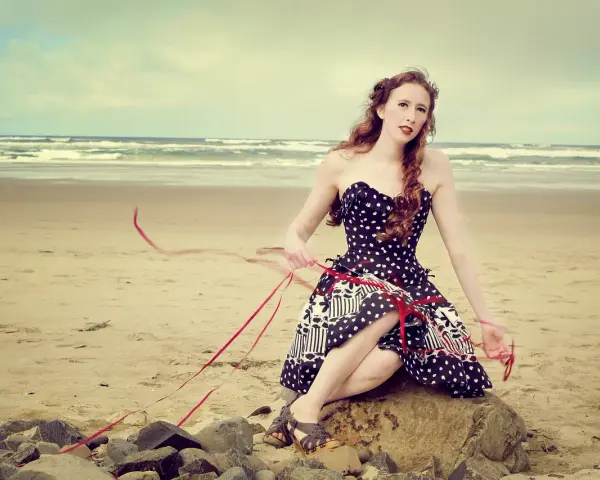 Jeune femme au style pin-up année 50, assise sur un rocher sur la plage, robe à pois, ruban rouge flottant au vent.