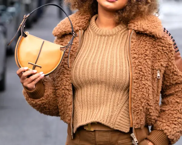 Une femme porte un pull beige côtelé, une veste en fausse fourrure marron et un sac à main jaune moutarde. Le jaune moutarde est une couleur qui va bien avec le beige.