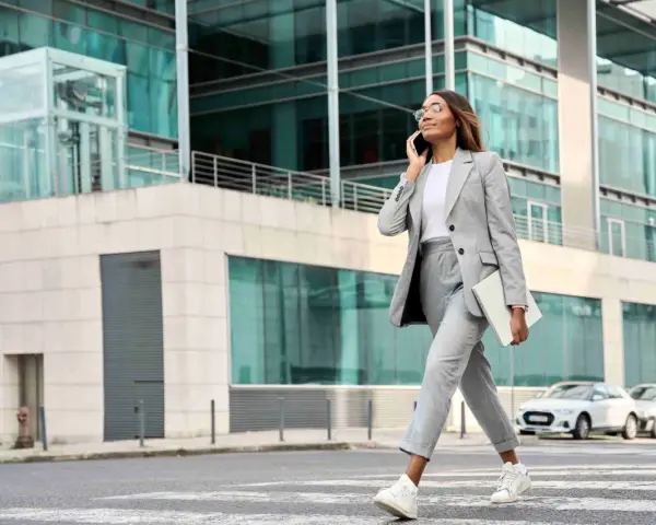 Femme en tenue d'entretien d'embauche d'été, tailleur gris, traversant la rue avec un ordinateur portable, parlant au téléphone.