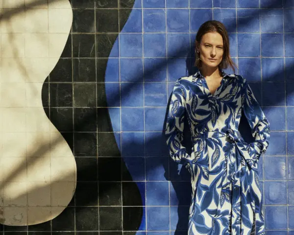 Une femme pose devant un mur de carreaux bleus et blancs. Sa robe à motifs bleus et blancs s'harmonise parfaitement avec le décor. Quelle couleur va bien avec le bleu vêtement ? Le blanc, évidemment !