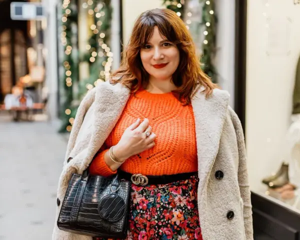 Une femme arbore une tenue avec pull orange vif, jupe fleurie et manteau douillet, prête pour une sortie festive.