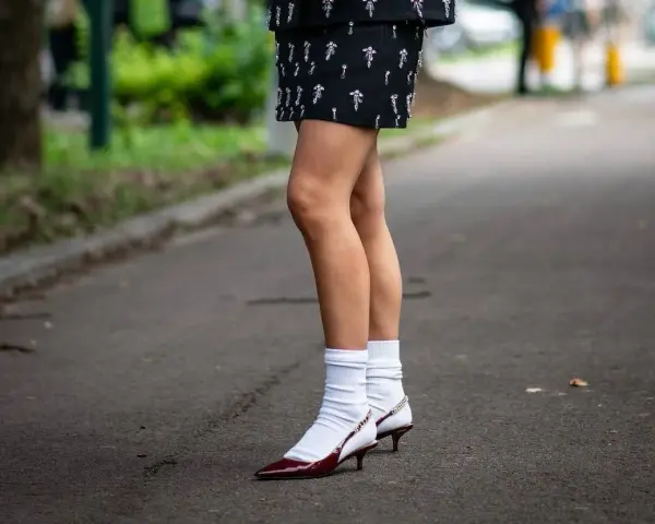 Jambes portant une jupe ornée et des escarpins bordeaux avec une chaussette par dessus chaussure blanche.