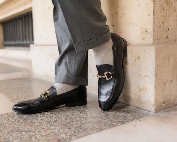 Un homme porte un pantalon gris retroussé et une chaussette mocassin noire avec une boucle dorée.