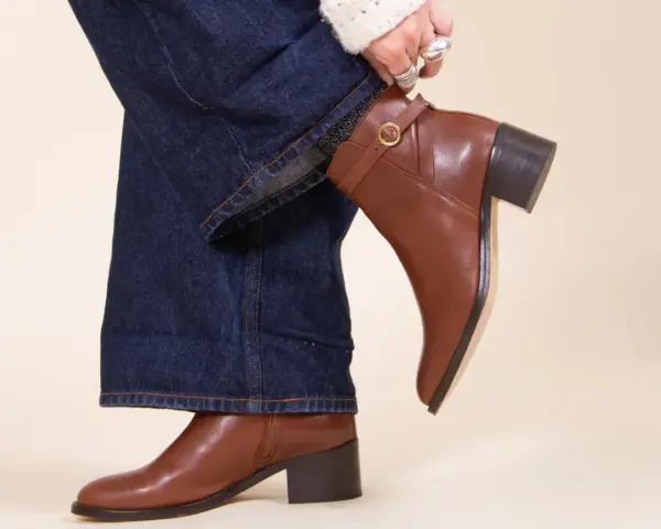 Une femme ajuste sa tenue avec bottines marron. Un jean évasé et un pull blanc complètent le look.